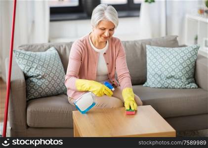 household and people concept - happy senior woman with detergent and sponge cleaning table at home. senior woman with detergent cleaning table at home
