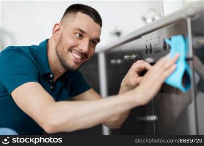 household and people concept - happy smiling man wiping table with cloth cleaning oven door at home kitchen. man with rag cleaning oven door at home kitchen. man with rag cleaning oven door at home kitchen
