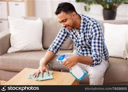 household and people concept - indian man with detergent cleaning table by rag at home. indian man cleaning table with detergent at home