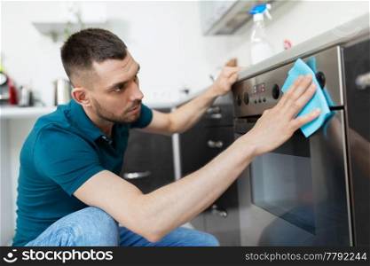 household and people concept - man wiping table with cloth cleaning oven door at home kitchen. man with rag cleaning oven door at home kitchen