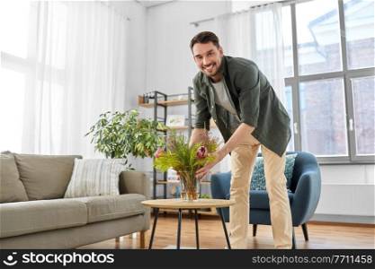 household, home improvement and interior concept - happy smiling young man placing flowers in vase on coffee table. man placing flowers on coffee table at home