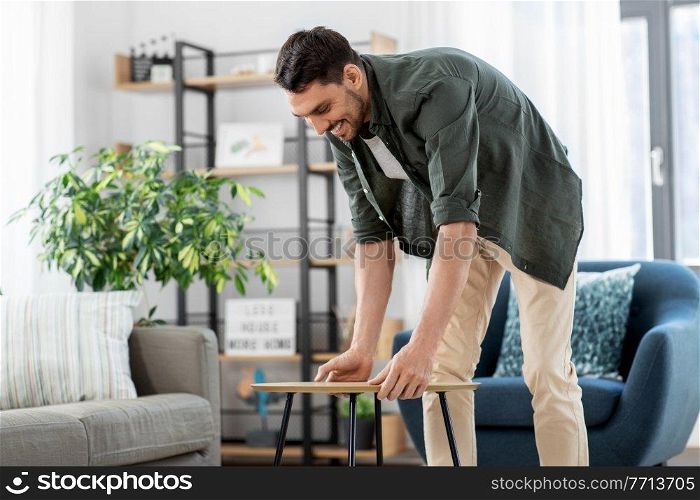 household, home improvement and interior concept - happy smiling young man placing coffee table next to sofa. man placing coffee table next to sofa at home