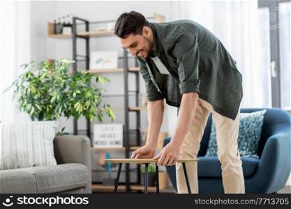 household, home improvement and interior concept - happy smiling young man placing coffee table next to sofa. man placing coffee table next to sofa at home