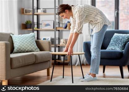 household, home improvement and interior concept - happy smiling young woman placing coffee table next to sofa. woman placing coffee table next to sofa at home