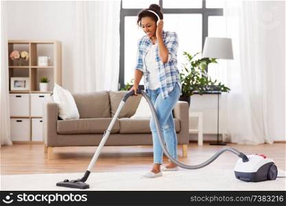 household, housework and cleaning concept - happy african american woman or housewife in headphones with vacuum cleaner at home. woman or housewife with vacuum cleaner at home