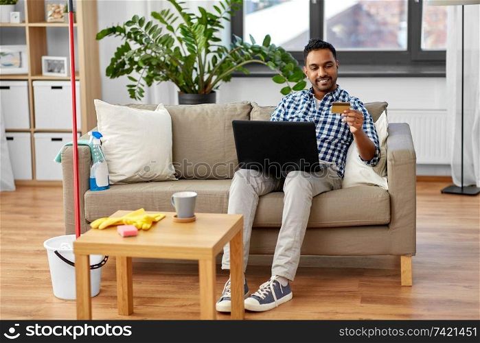 household, housework and technology concept - indian man with laptop computer and credit card shopping online after home cleaning. man with laptop shopping online after cleaning