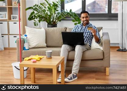 household, housework and technology concept - indian man with laptop computer and credit card shopping online after home cleaning. man with laptop shopping online after cleaning
