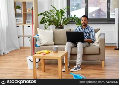 household, housework and technology concept - indian man with laptop computer, mop and bucket after home cleaning. man with laptop computer after home cleaning