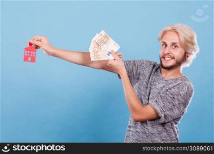 Household savings and finances, economy concept. Happy man holding money and keys to house, studio shot on blue background. Man holding money and keys to house
