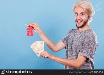 Household savings and finances, economy concept. Happy man holding money and keys to house, studio shot on blue background. Man holding money and keys to house