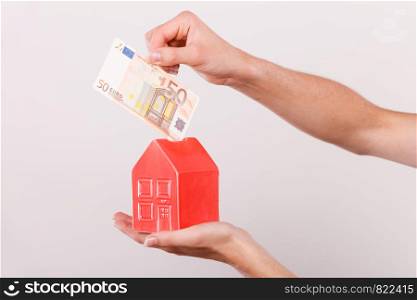 Household savings and finances, economy concept. Man putting euro money into a piggy bank in the shape of a house, studio shot on grey background. Man putting money into house piggybank