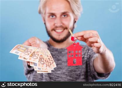 Household savings and finances, economy concept. Smiling man holding money and keys with small pendant in the shape of a house, studio shot on blue background. Man holding money and keys to house