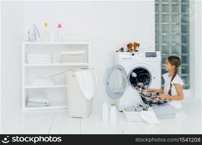 Housekeeping, children and domestic chores concept. Happy kid unloads washing machine, puts clean washed clothes in basin, curious dog looks from above, lies on washer in laundry room at home