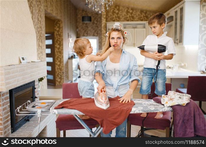 Housewife and kids playing with hair dryer at the ironing board. Woman with children doing housework at home together. Female person with daughter and son having fun in their house. Housewife and kids playing with hair dryer