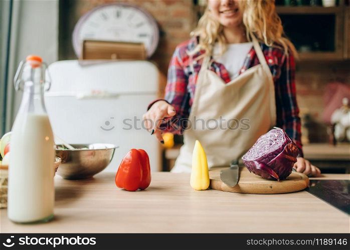 Housewife in an apron points finger at fresh pepper lying on wooden table, kitchen interior on background. Female cook making healthy vegetarian food, salad cooking. Housewife in apron points finger at fresh pepper