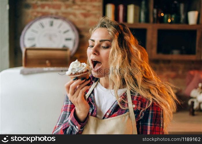 Housewife in apron eating sweet dessert with cream, kitchen interior on background. Female cook tastes fresh homemade cake. Chef eats pie. Housewife eating sweet dessert with cream