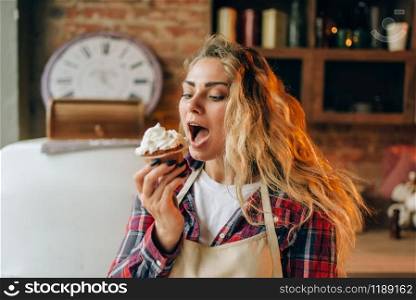 Housewife in apron eating sweet dessert with cream, kitchen interior on background. Female cook tastes fresh homemade cake. Chef eats pie. Housewife eating sweet dessert with cream