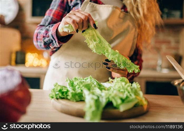 Housewife in apron prepares salad, kitchen interior on background. Female cook making healthy vegetarian food, vegetables preparation. Housewife in apron prepares salad on the kitchen