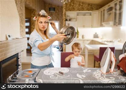 Housewife paints eyelashes at the ironing board, little kid looks at her. Woman with child at home together