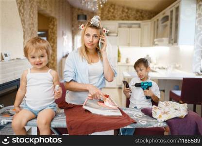Housewife talking by phone, kids fooling around on the kitchen. Woman with children playing at home together. Female person with daughter and son in their house