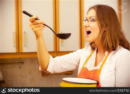 Housewife tasting soup meal dinner. Middle aged woman holding spoon ladle and pot. Housekeeper wearing orange apron preparing food.. Woman with ladle and pot in kitchen