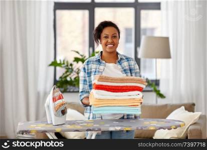 housework, laundry and housekeeping concept - happy african american woman with ironed linen and iron board at home. african american woman with ironed linen at home