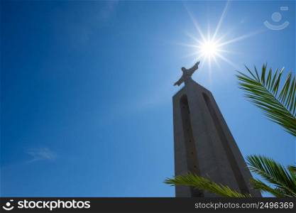 Huge statue of Christ the King through green palm leafs at clear summer sunny day in Lisbon Portugal with copy space