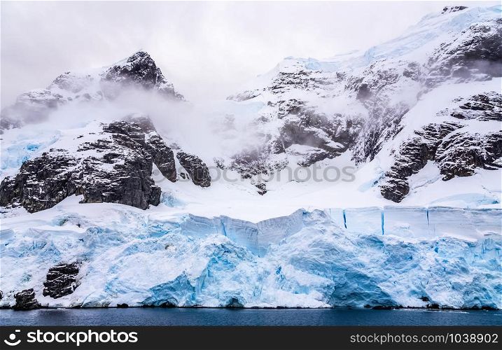 Huge steep stone rock covered with blue glacier and cloud with water in foreground, close to Argentine islands, Antarctic