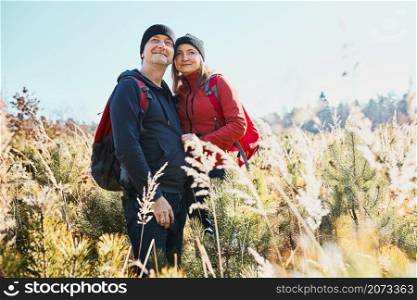Hugging couple enjoying nature while vacation trip. Hikers with backpacks looking at mountains view. People standing in tall grass on path to mountains on sunny day. Active leisure time close to nature