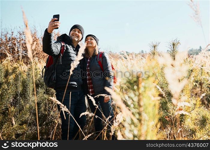 Hugging couple taking selfie while vacation trip. Hikers with backpacks on way to mountains. People walking through tall grass along path in meadow on sunny day. Active leisure time close to nature
