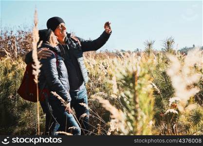 Hugging couple taking selfie while vacation trip. Hikers with backpacks on way to mountains. People walking through tall grass along path in meadow on sunny day. Active leisure time close to nature