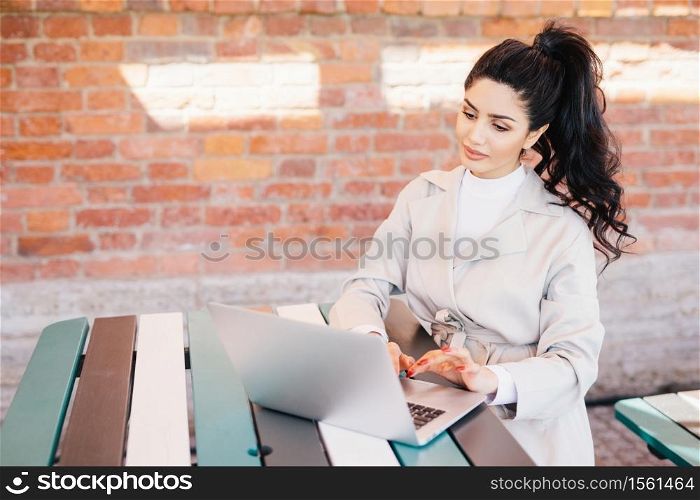 Human and technology. Young pretty woman with dark hair tied in pony tail dressed in white coat typing on laptop, texting friends via social networks sitting at table in cozy cafe over brick wall