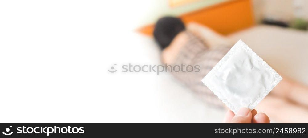 Human hand holding a packed condom against a blurred female background in white bed,horizontal copy space on white background