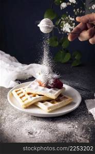 Human hand sprinkling powdered sugar on homemade waffles with strawberries and raspberries on white dish.