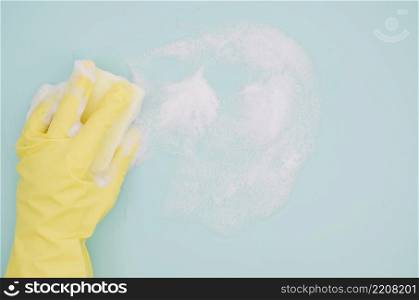 human hand wearing yellow gloves washing blue backdrop with soap sud