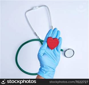 human hand with blue sterile gloves holding a red heart, white background with a stethoscope, top view
