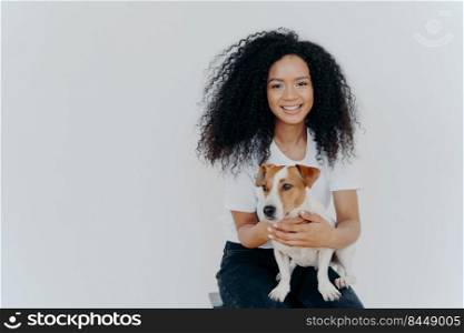 Humans and animals concept. Cheerful good looking woman with crisp hair, smiles pleasantly, plays with pedigree dog, sits on comfortable chair, makes memorable shot, pose against white background