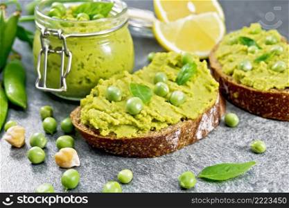 Hummus sandwiches with green peas and chickpeas, a jar of dip sauce, pea pods on a gray stone table background