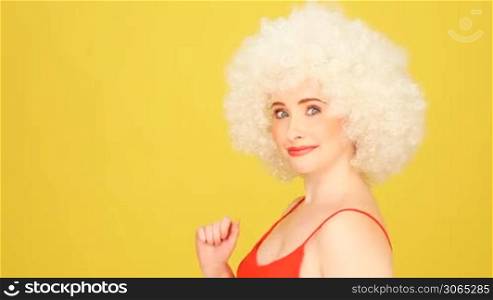 Humorous portrait of a young caucasian woman wearing a crazy white curly afro wig against a yellow studio background with copyspace.