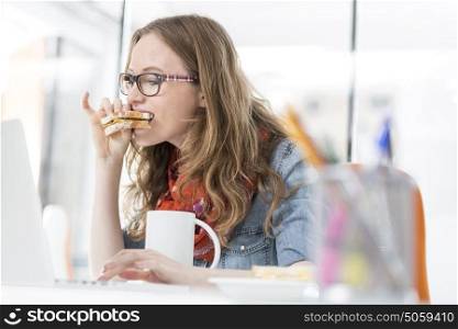 Hungry businesswoman eating sandwich while using laptop