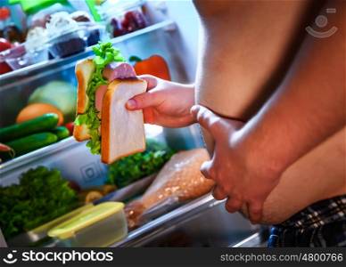 Hungry fat man holding a big sandwich in his hands and standing next to the open fridge. Unhealthy food.