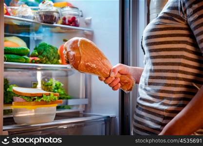 Hungry fat Woman holding a big turkey leg in his hands and standing next to the open fridge. Unhealthy food.
