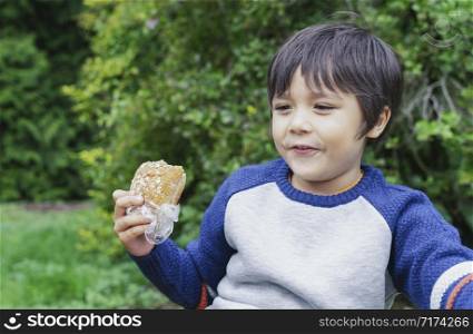 Hungry kid boy eating homemade bread sandwiches with mixed vegetables in the park, Child siting on green grass eating his snack picnic with blurry trees background, Spring outdoor activity