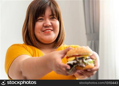 Hungry overweight woman smiling and holding hamburger and sitting in the bedroom, her very happy and enjoy to eat fast food. Concept of binge eating disorder (BED).