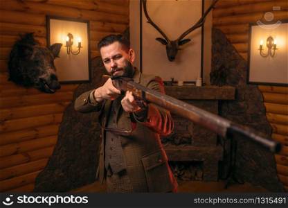Hunter man aims of the antique hunting rifle. Fireplace, stuffed wild animals, bear skin and other trophies on background