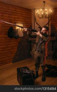 Hunter man aims of the antique hunting rifle. Fireplace, stuffed wild animals, bear skin and other trophies on background. Hunter man aims of the antique hunting rifle
