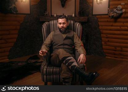 Hunter man in traditional british clothing sitting in a chair after hunting and drink whiskey. Fireplace, stuffed wild animals, bear skin and other trophies on background. Hunt lifestyle. Hunter man sitting in a chair and drink whiskey