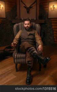 Hunter man in traditional british clothing sitting in a chair after hunting and drink whiskey. Fireplace, stuffed wild animals, bear skin and other trophies on background. Hunt lifestyle