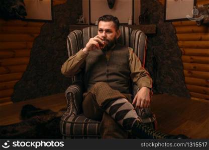 Hunter man in traditional vintage hunting clothing drink luxury alcohol after successful hunt. Fireplace, stuffed wild animals, bear skin and other trophies on background. Hunter man drink alcohol after successful hunt