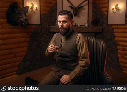Hunter man in traditional vintage hunting clothing drink luxury alcohol after successful hunt. Fireplace, stuffed wild animals, bear skin and other trophies on background. Hunter man drink alcohol after successful hunt